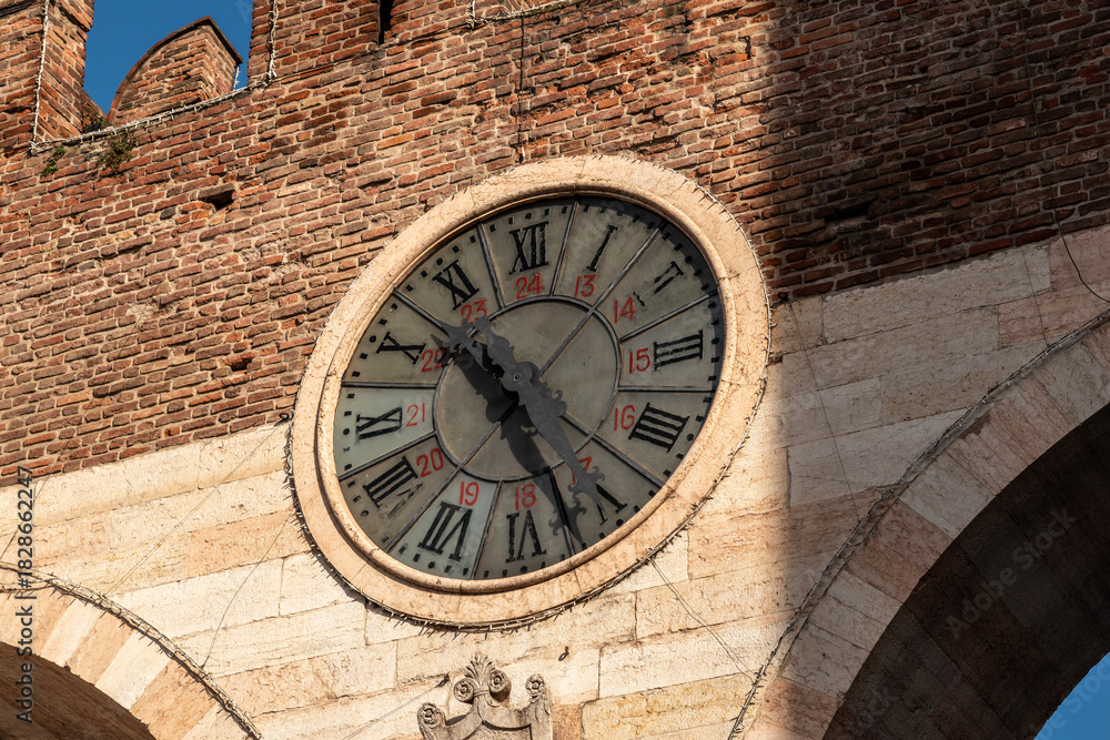 Fototapeta premium Close-up view of the large historic clock on the medieval city gate Portoni della Bra in Verona, Italy. The round clock with Roman numerals and 24-hour markings is set in a brick and stone wall with a