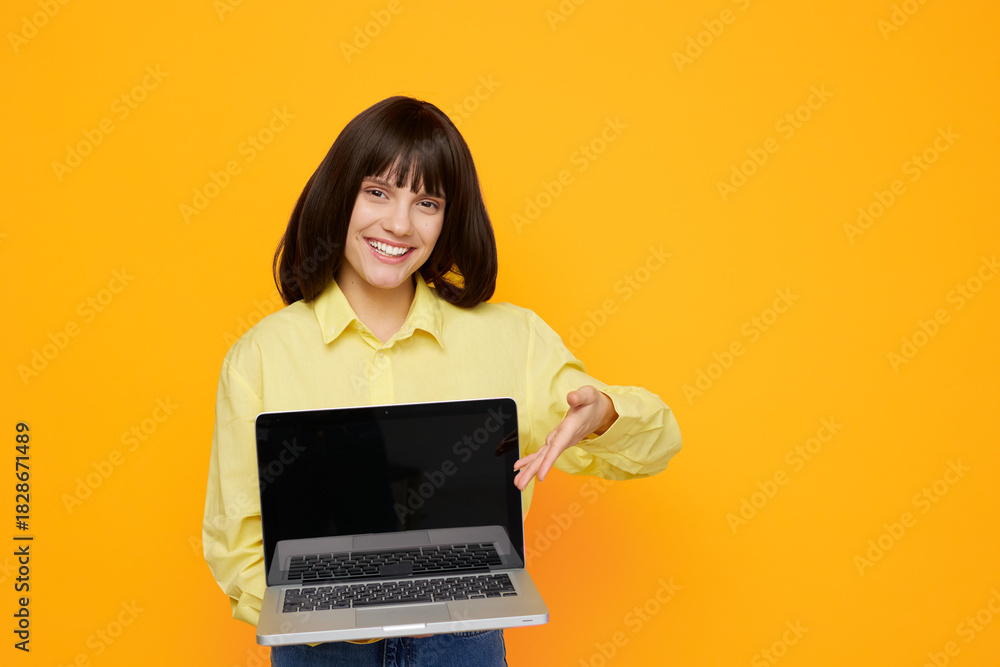 Fototapeta premium A cheerful woman stands against a vivid orange backdrop, proudly presenting a laptop. This image conveys online learning, remote work, and modern technology with approachable energy.