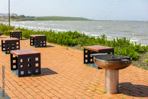 Fototapeta Naklejka Na Ścianę i Meble -  Sunny summer day at Sillamae beach promenade (Estonian - Sillamäe), Estonia. Modern brick-paved area with steel barbecue grill and wooden benches by the Baltic Sea. 