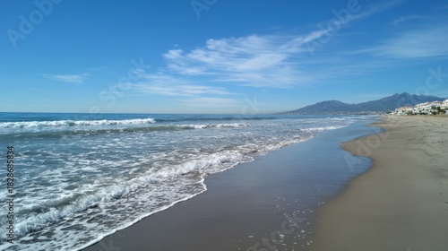 Clear skies and soft waves create a peaceful scene at the beach. The shoreline features smooth sand and inviting water with mountains visible in the distance.
