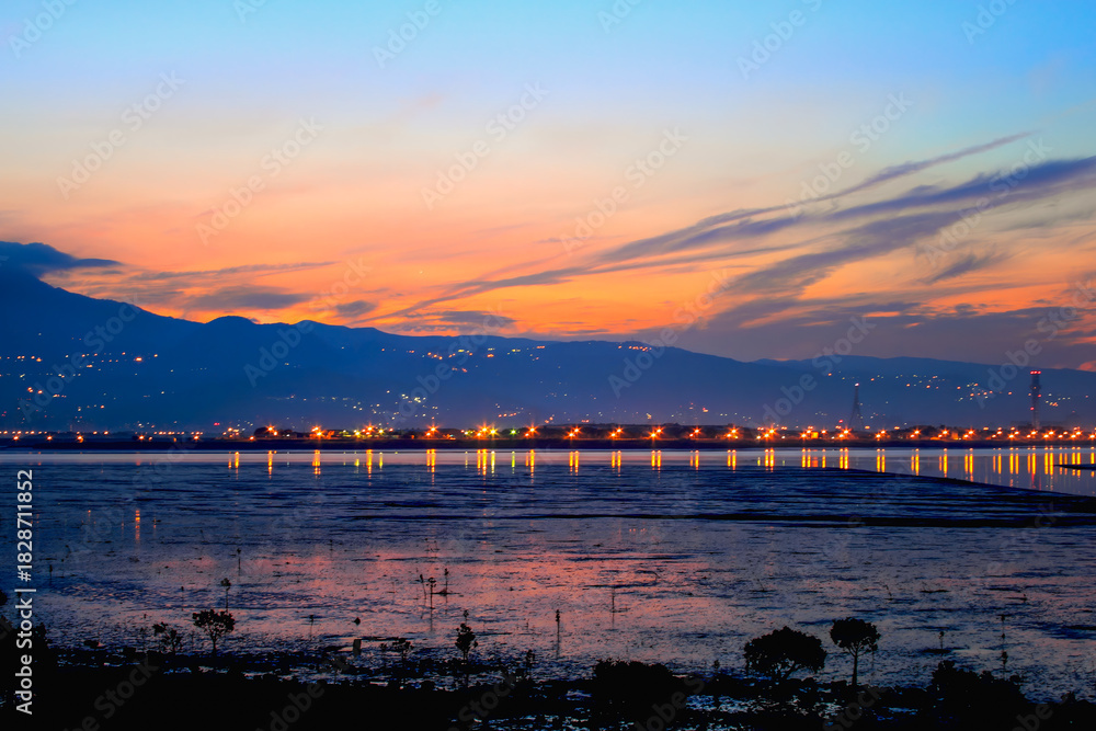 Obraz premium Stunning twilight scenery over Wugu Wetland in New Taipei, Taiwan. Mountains silhouetted against golden sky, city lights reflecting on calm waters.