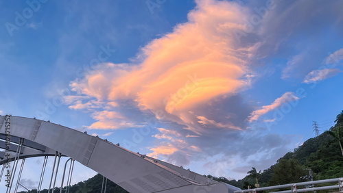 Unique lenticular cloud formation illuminated with warm orange light above the modern bridge structure in Wulai New Taipei City Taiwan during dawn.