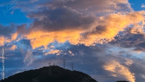 Stunning sky over Wulai New Taipei City Taiwan at dawn or dusk. Dramatic orange and gray clouds hover above power transmission towers on a forested mountain.