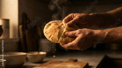 Male hands breaking fresh hot bread with steam. Rustic homemade loaf with falling crumbs in dark kitchen