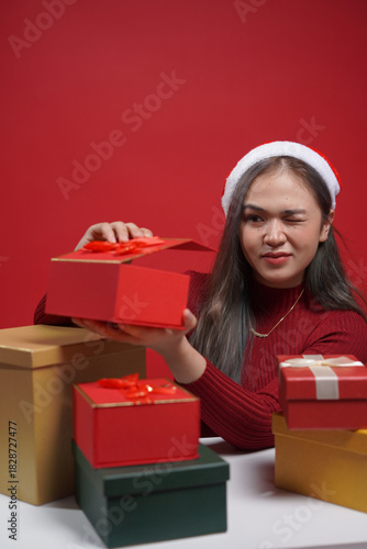 Beautiful woman in Christmas outfit presenting gift box with cheerful expression – red studio background, holiday promotion and winter celebration