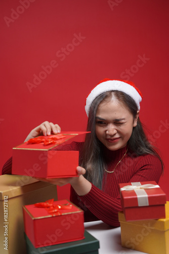 Beautiful woman in Christmas outfit presenting gift box with cheerful expression – red studio background, holiday promotion and winter celebration