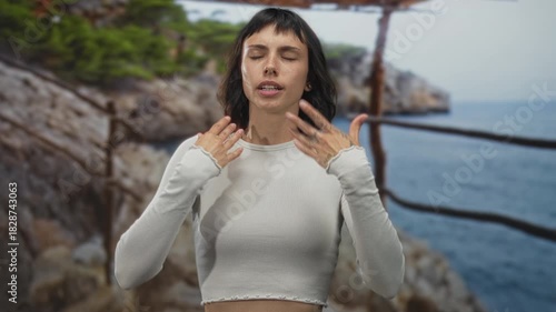 Young hispanic woman fans face with hands beside a rugged rocky coast railing in forest; relief from heat.