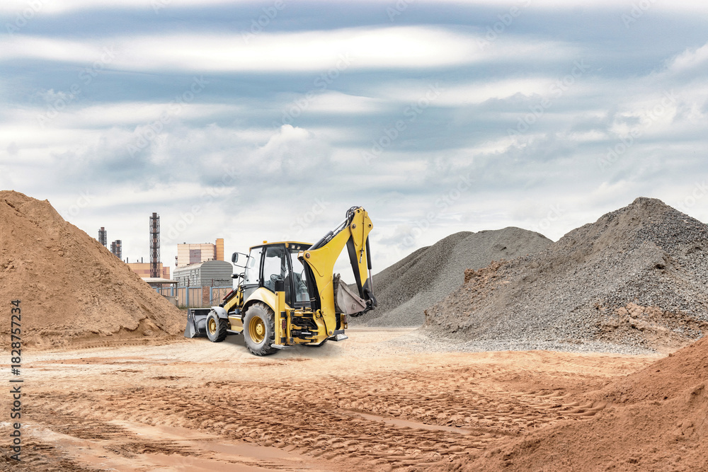 Obraz premium A yellow backhoe works on a construction site, digging through mounds of dirt and gravel with industrial buildings in the background on a cloudy day