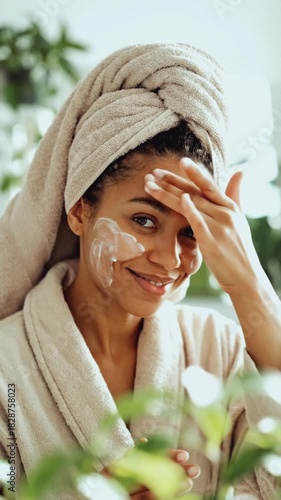 Vertical video of a happy woman applying face cream during her skincare routine. Young female in a bathrobe and towel smiling while massaging moisturizer on her face. Beauty and self-care concept