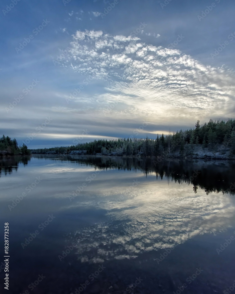 Fototapeta premium Calm lake reflecting evening clouds and forest shoreline under soft fading blue light