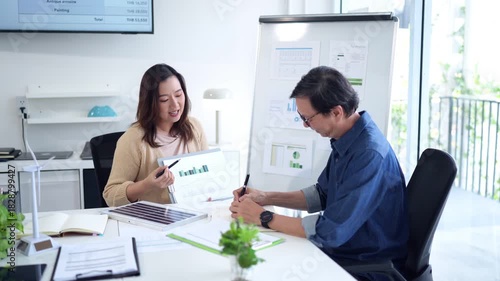 Asian business professional woman presents financial data to older man discussing sustainable energy strategies office meeting productivity