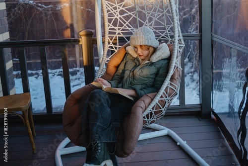 A 12-year-old teenage girl reads a book on a cozy winter porch. Authentic moment of quiet relaxation, literary escape and seasonal tranquility