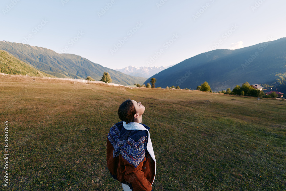 Fototapeta premium Woman in coat stands in a meadow field looking up at sky and distant mountains, serene landscape and travel concept with open space, solitude and nature scenery.