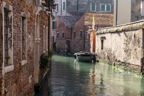 Narrow Venice backstreet canal lined with weathered brick walls, mossy foundations and small working boat creating authentic rustic lagoon mood.