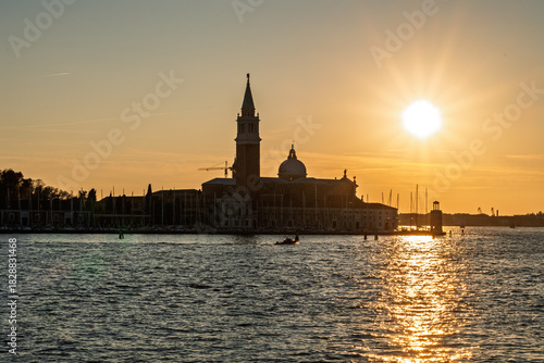 Golden sunset over San Giorgio Maggiore island in Venice with warm sky, church silhouette and sparkling light trail reflected on lagoon waves.