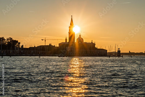Dramatic sunburst setting directly behind San Giorgio Maggiore bell tower in Venice, casting radiant rays and long shimmering reflection across lagoon.