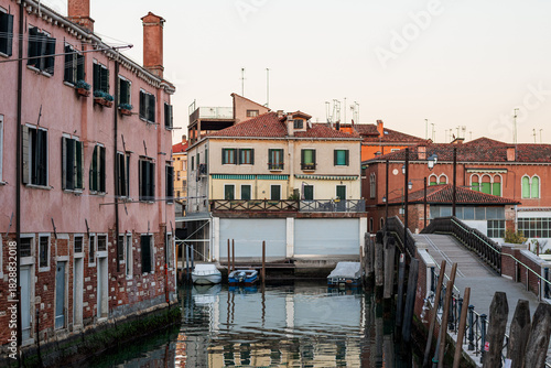 Charming Castello waterfront in Venice with pastel residential buildings, small boats docked by shuttered garage doors and pedestrian bridge crossing the canal.