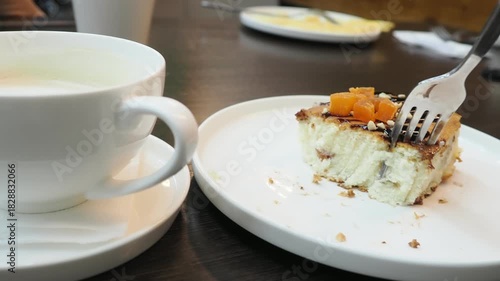 On the table of a cozy cafe there is a cup of aromatic cappuccino with creamy foam and a delicious dessert. A girl is eating it with a fork. In the background, people are looking at their phone and sc