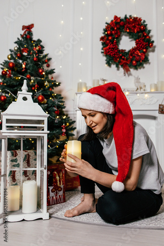 Woman wearing a Santa hat enjoys a candle by a decorated Christmas tree in a cozy holiday setting