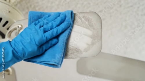 A gloved hand with a blue cloth cleans a dusty ceiling fan blade with a light-colored blade