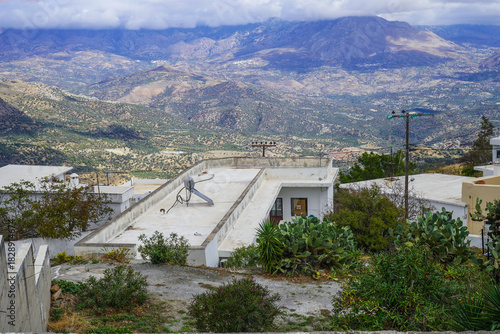 Fototapeta Naklejka Na Ścianę i Meble -  View from a village street in Melampes, Crete, looking across a low rooftop equipped with a solar heater and TV antenna, toward the dramatic backdrop of the rugged Cretan mountains.
