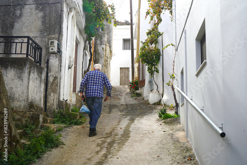 Fototapeta Naklejka Na Ścianę i Meble -  An old man in a blue plaid shirt walks up a steep, narrow alley in Melampes, Crete, carrying a light blue bag. The path is lined with white and weathered walls and climbing vines.
