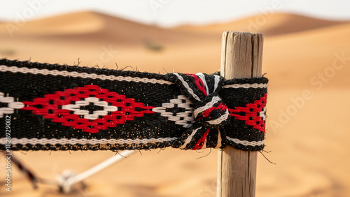 Macro close-up of traditional black and red Sadu weaving strap tied to wooden stake in Saudi desert with sand dunes background