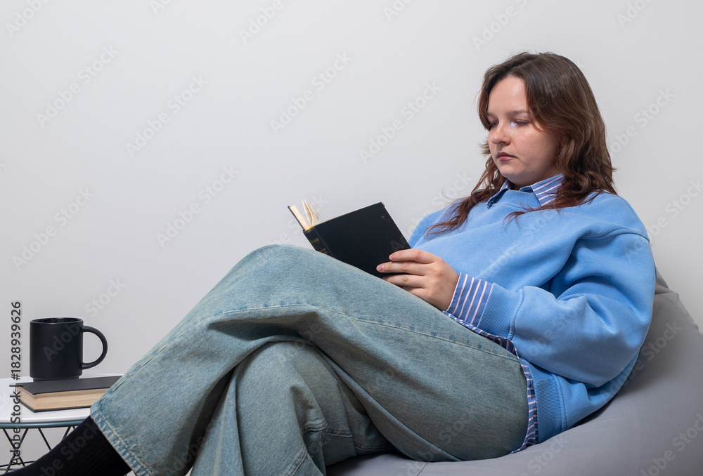 Obraz premium Young woman relaxing at home, sitting comfortably with legs crossed in a soft chair while reading a