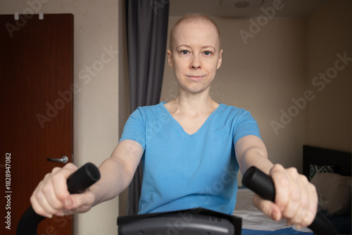 Focused bald woman pedaling on exercise bicycle, cancer treatment side effects and safe home workouts, low impact training for circulation and lungs, rehabilitation