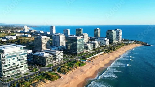 Modern coastal city skyline with high-rise buildings overlooking a sandy beach and ocean waves under a clear blue sky.