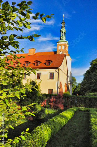 Zabytkowy kościół z czerwonymi dachami i wieżą otoczony zielonymi ogrodami pod błękitnym niebem. Lubiń - Klasztor Opactwa Benedyktynów.