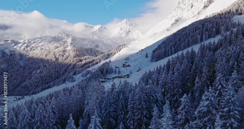 Aerial footage of snowy alpine peaks in Austria with a glacier rising through morning mist. Blue sky, sunlight, and conifer forests in the foreground create a peaceful winter atmosphere.