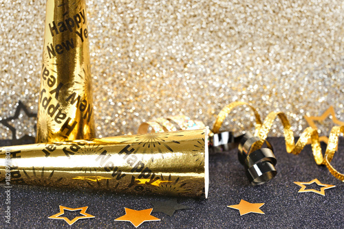 New Years Eve party decor on a twinkling gold and black background. Noisemakers, ribbon, confetti.