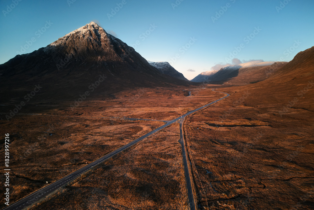 Naklejka premium Aerial view of Buachaille Etive Mor and the road through Glen Coe, Scotland. Dramatic mountain landscape, warm evening light, and vast moorland scenery in autumn. Natural landscape, no people