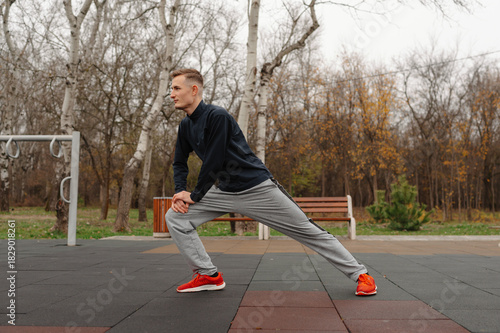 caucasian man lunging stretch outdoors park, wearing grey tracksuit and red shoes, focused warmup before run,