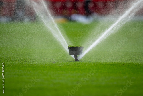 Close-up of an automatic sprinkler watering a vibrant green grass field, with water droplets spraying outwards. Ideal for themes of maintenance, growth, sports, or agriculture.