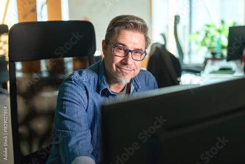 Businessman working with computer in office. Business lifestyle - casual middle aged man sitting at desk in front of screen, smiling, happy.