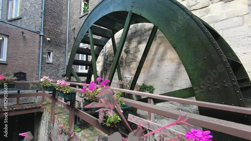 Old water wheel turning beside historic stone buildings, surrounded by blooming flowers. Rustic architecture and traditional engineering create a charming, vintage European atmosphere