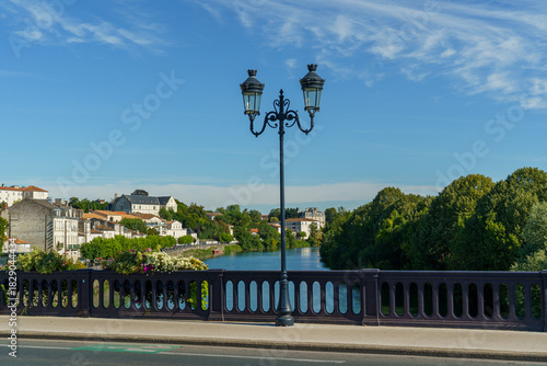 Fototapeta Naklejka Na Ścianę i Meble -  A street with a bridge and a river La Charente in city Saintes France. The street is lined with trees and houses. The sky is blue and clear