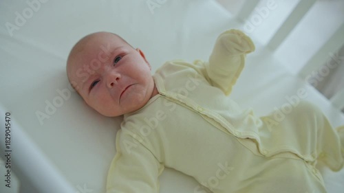 A one-month-old baby is lying in a white wooden crib, wearing a yellow onesie, kicking its legs and crying