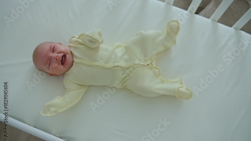 A one-month-old baby is lying in a white wooden crib, wearing a yellow onesie, kicking its legs and crying