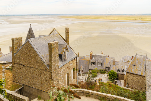 Several buildings with ancient stone tile roofs photographed from above at the Chateau de Mont Saint-Michel in Normandy, France Sand and the Atlantic Ocean are visible in the background Evening time