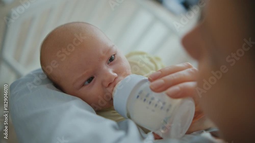 A mother is feeding a small baby formula from a bottle in front of a crib close up. The baby is eating with appetite and looking at her