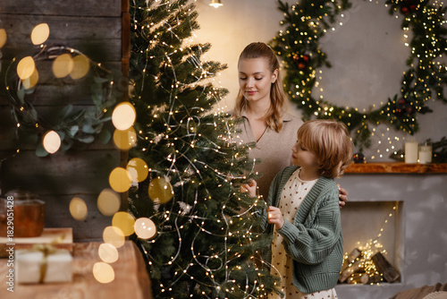Caucasian mother and daughter, blonde, decorating Christmas tree together, standing close, front view, cozy living room with warm lights and wreaths. Concepts family, holiday, togetherness, Christmas.