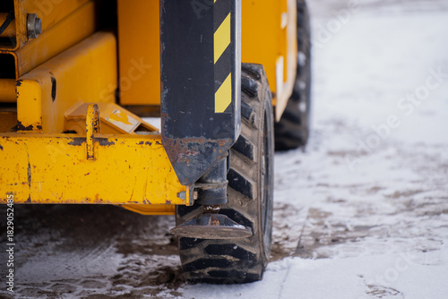 Snowy worksite scene featuring a yellow construction machine with visible tire, metal frame, worn paint and warning stripes indicating industrial winter conditions