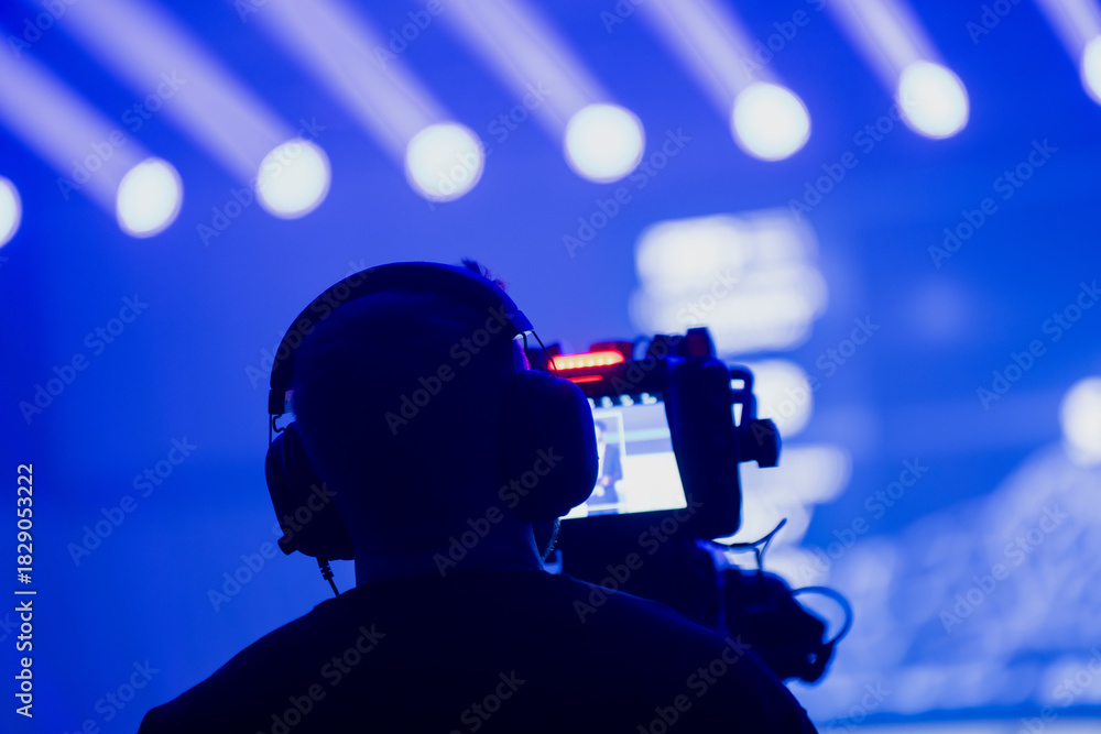 Fototapeta premium Camera operator filming a live event under intense blue stage lighting with bright beam patterns, showcasing professional broadcast and event production workflow