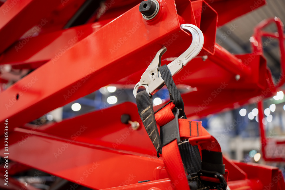 Naklejka premium Safety harness with metal carabiner clipped to red industrial lift structure, highlighting fall protection equipment used in construction and maintenance work