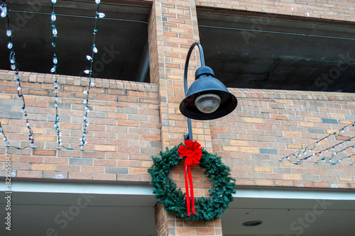 Classic green Christmas wreath with a red bow hanging on a brick wall beneath an industrial gooseneck lamp fixture during the holiday season
