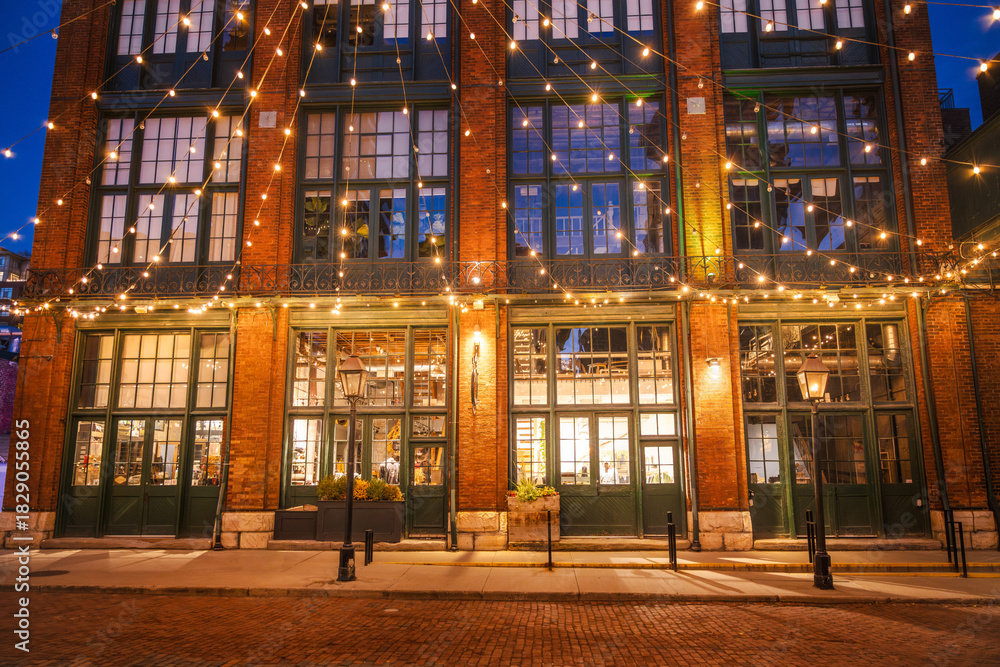 Obraz premium Historic brick building decorated with warm string lights in the Distillery District of Toronto during the evening.