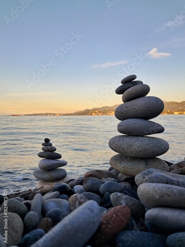 Two carefully balanced stacks of smooth, grey stones (cairns) stand on a pebble beach by the calm sea, symbolizing balance, meditation, or tranquility.